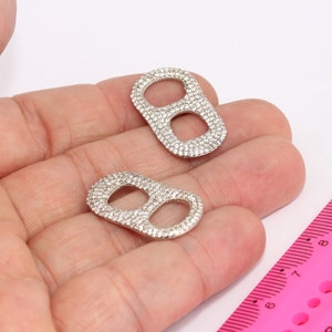 Könnte beinhalten: Zwei silberfarbene Metallringe mit einem Doppel-Schleifendesign, jeder Ring ist mit kleinen, klaren Strasssteinen besetzt.