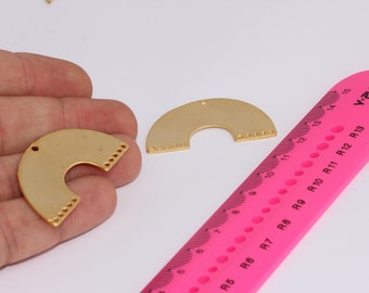 May include: Two gold-colored metal half-circle shapes with holes around the edges. The shapes are about 2 inches in diameter. A pink ruler with black markings is in the background.