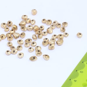 Könnte beinhalten: Eine Sammlung goldfarbener Metallperlen, verstreut auf einer weißen Oberfläche. Die Perlen sind klein und haben eine abgerundete, unregelmäßige Form. Ein grünes Lineal ist im Hintergrund sichtbar.