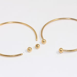 Könnte beinhalten: Zwei goldene Armreifen mit einer Kugel an jedem Ende. Ein Armreif ist offen und der andere ist geschlossen.