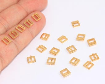 May include: A group of gold-colored metal square buckles, some are laying flat and others are standing upright on a white surface.