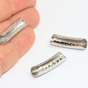 May include: Three silver-colored, hammered-texture metal tube beads. The beads are cylindrical with slightly curved sides and open ends. One bead is held in a hand, while two others are on a white surface. These beads are likely for jewelry making.