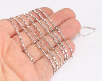 May include: A silver chain necklace with a flat, rectangular link design. The chain is shown in a hand, with multiple strands of the chain layered on top of each other.