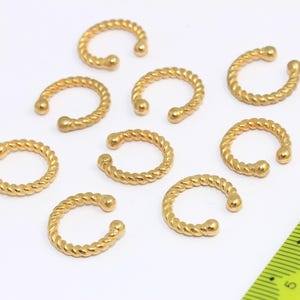 Puede incluir: Un conjunto de puños de oreja dorados con un diseño de cuerda trenzada. Los puños de oreja están dispuestos sobre una superficie blanca, con una regla amarilla en el fondo. Los puños de oreja tienen forma curva y están abiertos por un extremo.