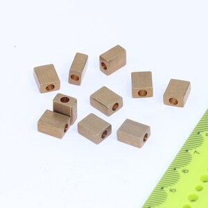 May include: A group of ten gold-colored metal beads with a hole through the center of each bead. The beads are arranged in a random pattern on a white surface. A green ruler is visible in the bottom right corner of the image.
