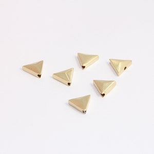 Peut inclure: Six petites perles triangulaires en métal doré sont dispersées sur une surface blanche. Chaque perle a un petit trou sur un côté. Les perles ont une finition mate et sont espacées uniformément.