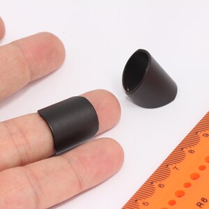 Puede incluir: Dos protectores de dedos negros, uno en un dedo y otro acostado sobre una superficie blanca. Una regla naranja brillante con marcas negras está en el fondo.