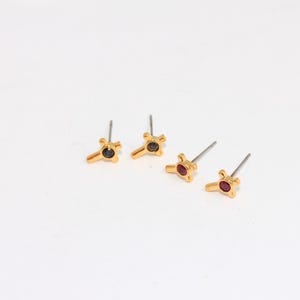 May include: Three pairs of gold stud earrings with a small, geometric design. One pair has a black stone, one pair has a red stone, and one pair has a purple stone.