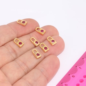 May include: Eight gold-colored padlock charms with a pink rhinestone in the center. The charms are arranged on a hand, with a pink ruler in the background.