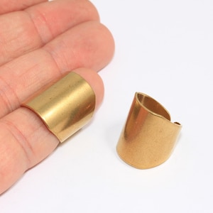 May include: Two gold-coloured, open-ended rings. One ring is on a finger, the other is separate. The rings have a simple, curved design. The background is plain white, highlighting the jewellery's metallic sheen. The rings appear to be made of brass or a similar metal.