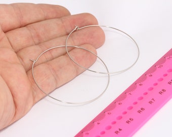 May include: Two silver hoop earrings, one slightly larger than the other, are shown on a hand against a white background. A pink ruler is partially visible in the bottom right corner of the image.