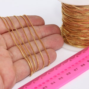 Puede incluir: Un carrete de cadena de color dorado, con varios hilos de cadena dispuestos sobre una mano, junto a una regla rosa marcada con números y la letra "R".