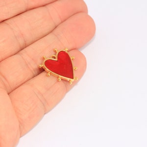 May include: A gold heart charm with a red enamel center. The charm has small gold beads around the edge.