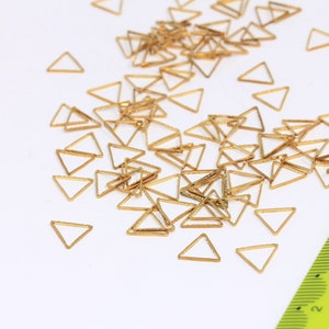 Pode incluir: Uma coleção de pequenos pingentes triangulares dourados espalhados sobre uma superfície branca. Uma régua verde é parcialmente visível. Os pingentes são delicados e parecem ser de metal, possivelmente para fazer joias ou artesanato.