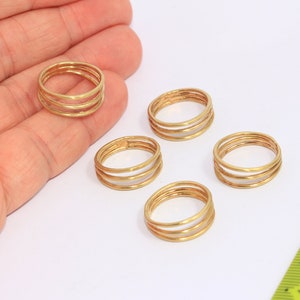May include: Five gold-toned triple-band rings, each with a slightly different diameter, are arranged on a white surface. A yellow ruler is visible in the background.