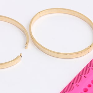 Op de afbeelding: Twee gouden armbanden, een open en een gesloten, met een roze liniaal op de achtergrond.