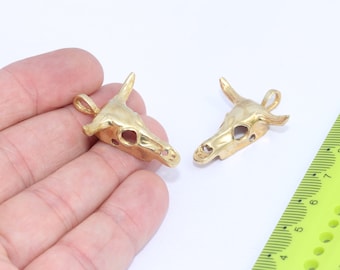 May include: Two gold-colored bull skull pendants with a loop for attaching a chain. The pendants are on a white surface.