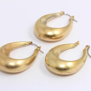 May include: A pair of gold hoop earrings with a smooth, rounded shape. The earrings are made of metal and have a brushed finish.