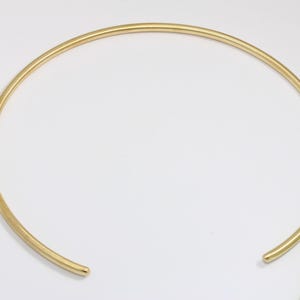 Peut inclure: Un collier ras du cou doré de conception simple et minimaliste. Le collier est un cercle lisse et ininterrompu, ouvert à l'avant. Une règle verte et jaune est en arrière-plan, fournissant une échelle. Le collier est probablement en métal.