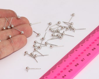 May include: A close-up of a pile of silver earring posts with round balls on the ends. The earring posts are scattered on a white surface next to a pink ruler with black markings.