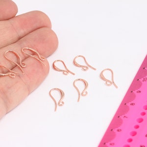 May include: Seven rose gold earring hooks, also known as ear wires, are displayed on a white surface. A pink ruler with black markings is in the background.