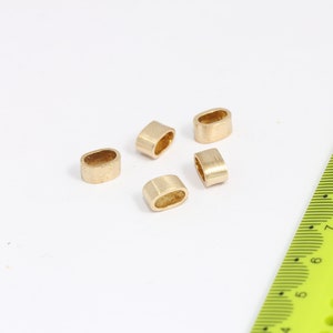 May include: Five small, gold-colored, oval-shaped metal beads are scattered on a white surface. A yellow ruler with black markings is partially visible in the frame, providing scale. The beads appear to be components for jewelry making.
