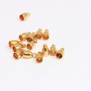May include: A group of 14 gold-coloured metal end caps for cords or jewellery making. The caps are small and have a rounded shape.