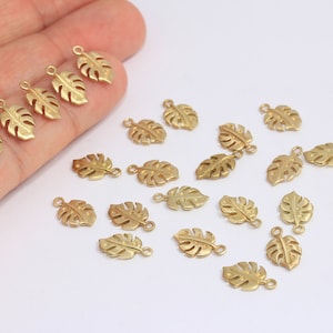 May include: A collection of 20 gold-toned metal charms shaped like monstera leaves. The charms have a hole at the top for attaching to jewellery or other crafts.