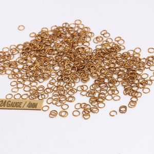 Op de afbeelding: Een grote stapel gouden springringen, 24 gauge en 4 mm groot. De springringen zijn verspreid over een wit oppervlak. Een klein label luidt "24 Gauge / 4mm".