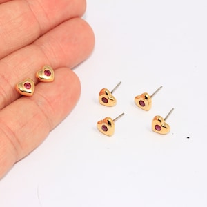 May include: Four gold heart-shaped earrings with a small red gemstone in the center. The earrings have a post back.