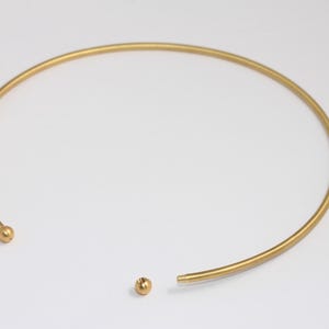 Könnte beinhalten: Eine goldfarbene Metall-Choker-Kette mit schlichtem, minimalistischem Design. Die Kette ist offen und hat an den Enden zwei kleine runde Kugeln.