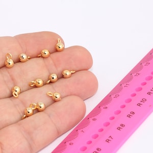 May include: A close-up of a hand holding a group of small, gold-coloured metal beads with loops. The beads are approximately 6mm in diameter. A pink ruler with black markings is in the background.