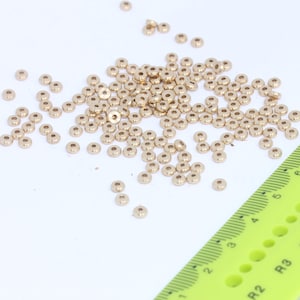 May include: A large pile of gold-colored metal beads, scattered on a white surface. A green ruler with black markings is visible in the bottom right corner of the image.