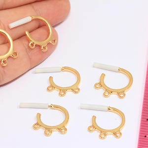 May include: Gold-colored earring components with a curved design and three small loops at the bottom. Each component has a white, rectangular post. The components are displayed on a white surface.