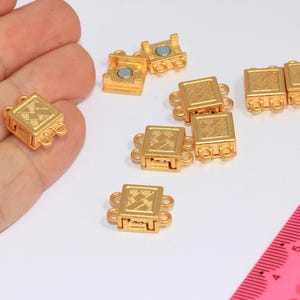 May include: Gold-colored magnetic clasps with a square design and decorative detailing. The clasps have loops on each side for attaching to jewelry. A pink ruler is in the background.