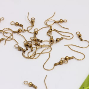 Puede incluir: Primer plano de un montón de ganchos para pendientes de color dorado. Los ganchos son de metal y tienen un pequeño bucle en la parte superior y una pequeña bola en la parte inferior.