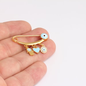 May include: Gold-colored safety pin with three charms: a Hamsa hand, a light blue heart, and a circular charm with a blue eye design. The pin has a light blue eye design at the top.