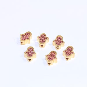 Puede incluir: Seis amuletos de mano de Hamsa de color dorado con diamantes de imitación rosas. Los amuletos están esparcidos sobre una superficie blanca.
