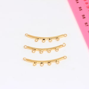 May include: Three gold-coloured, curved jewellery connectors with multiple loops for attaching beads or other components. The connectors are arranged on a white surface, with a pink ruler in the background.