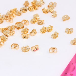 Può includere: Una collezione di chiusure per orecchini color oro sparse su una superficie bianca. Le chiusure sono piccole, con un design a forma di farfalla. Un righello rosa è visibile nell'angolo in basso a destra, che fornisce la scala. Le chiusure sono per la creazione di gioielli.