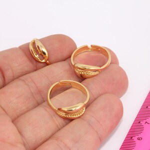 May include: Three gold-colored rings with a seashell design. The rings are on a hand, and a pink ruler is in the background.