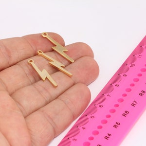 May include: Three gold-colored lightning bolt charms with a hole at the top for attaching to jewelry or other crafts. The charms are shown on a hand against a pink ruler.