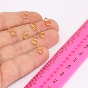 Op de afbeelding: Een close-up van een hand die verschillende gouden dubbele lus springringen vasthoudt. De ringen zijn ongeveer 1,25 cm in diameter. Een roze liniaal is op de achtergrond te zien.