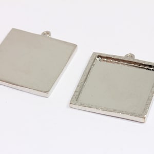 Puede incluir: Dos colgantes cuadrados plateados con un lazo para sujetar una cadena. Uno es un cuadrado macizo, y el otro tiene una textura martillada alrededor de los bordes y un área empotrada para agregar un diseño. Los colgantes están sobre un fondo blanco.