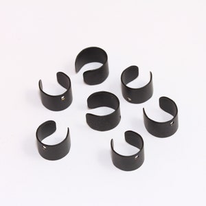 Puede incluir: Ocho anillos de metal negro con un pequeño agujero en cada uno. Los anillos están dispuestos en círculo sobre un fondo blanco.