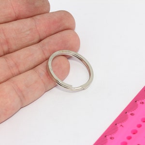 Puede incluir: Un llavero plateado con un diámetro de aproximadamente 3,8 cm. El llavero se muestra sobre una superficie blanca junto a una regla rosa.