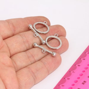 Pode incluir: Dois fechos de correr de metal prateado com bolas redondas em cada extremidade. Os fechos estão abertos e prontos para serem presos a um colar ou pulseira.