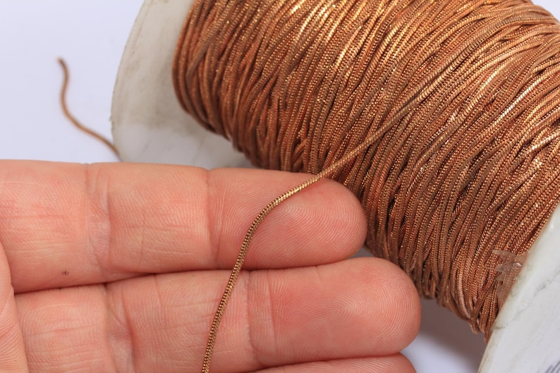 May include: Close-up of a spool of copper-colored metallic thread. The thread is a shiny, thin, and flexible material, ideal for jewelry making or crafts. The spool is white and partially visible, with the thread wound tightly around it.