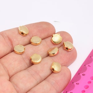 Könnte beinhalten: Nahaufnahme einer Hand, die acht goldfarbene, muschelförmige Perlen hält. Die Perlen haben einen Durchmesser von etwa 1 cm und eine glatte, glänzende Oberfläche.