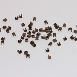 May include: A scattering of small, bronze-coloured metal crimp beads on a white surface. These C-shaped beads are used in jewellery making and crafting to secure beads and other components.